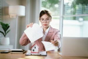 A young real estate agent in a pink blazer tears up a contract at her desk, with a small model house placed in front of her.