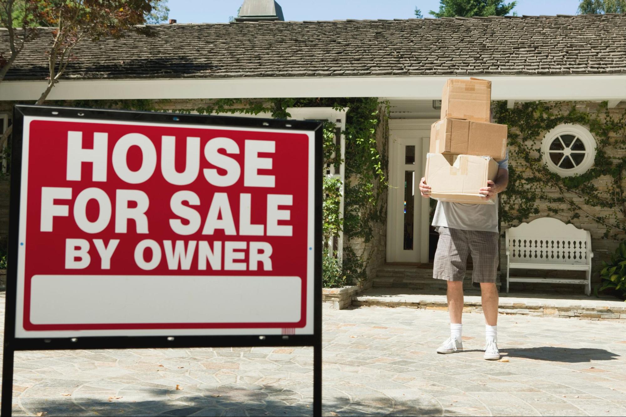 For sale by owner sign outside house with homeowner carrying stack of boxes in yard