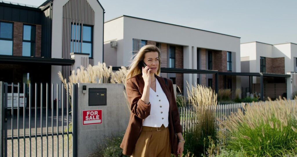 female real estate agent talking on the phone in front of a house condo with a for sale sign behind