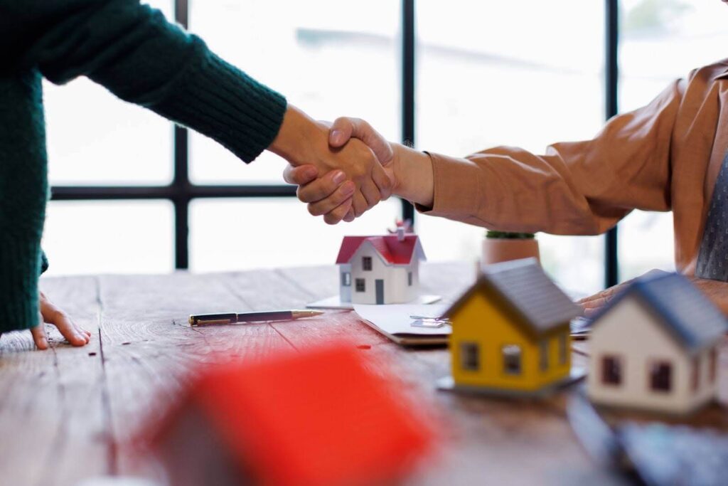a close-up on a real estate agent shaking the hand of the home seller above the table with small house miniatures on top of it